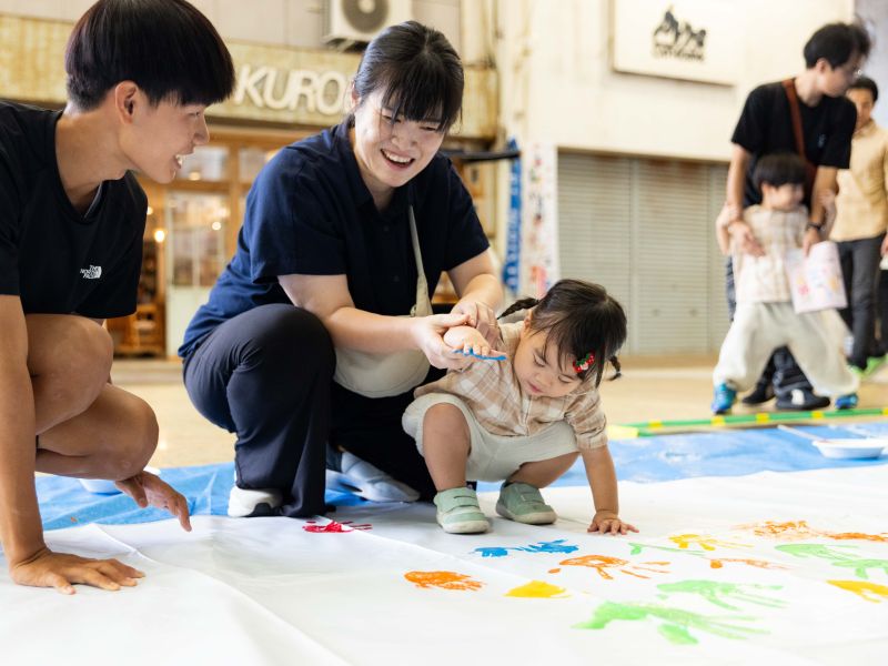 手にインクを塗って看板にスタンプする幼児と、サポートする大人の写真